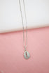 A delicate silver box chain necklace features a sparkling pavé-set cowrie shell pendant displayed against a soft pink and white background.