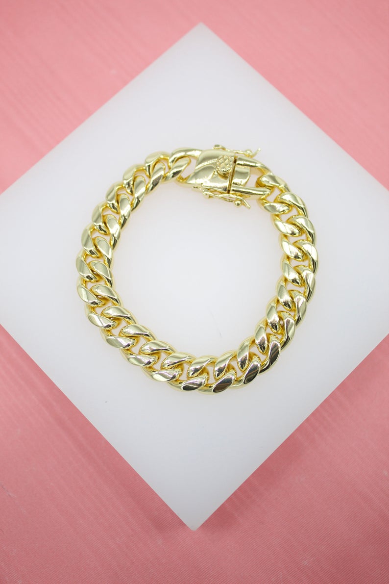 A thick, polished gold-plated Miami Cuban link chain bracelet rests on a white display stand against a pink background.