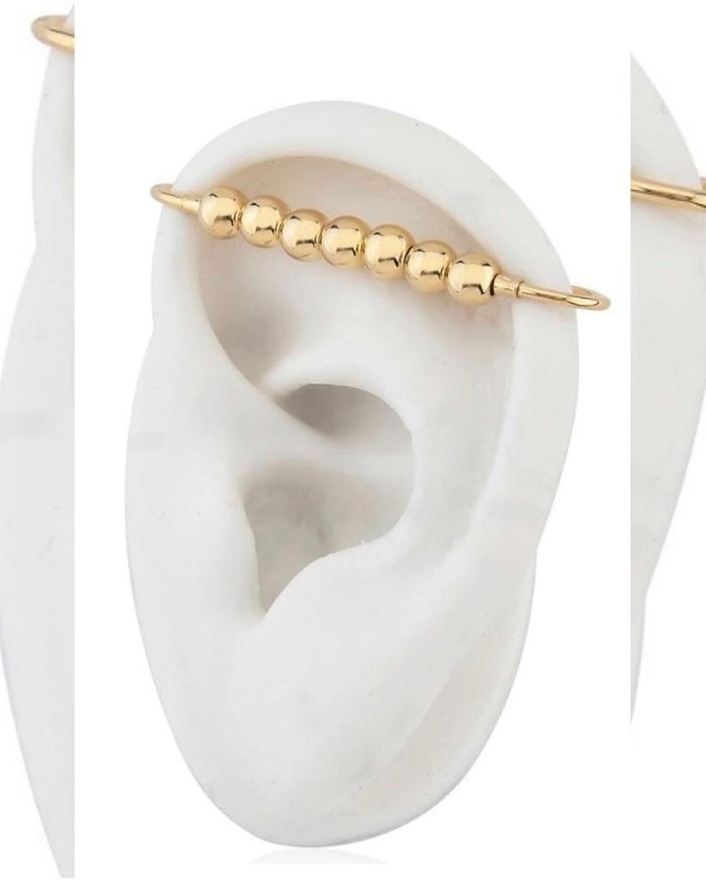A gold industrial barbell earring featuring a row of smooth metal beads is showcased on a white model ear.