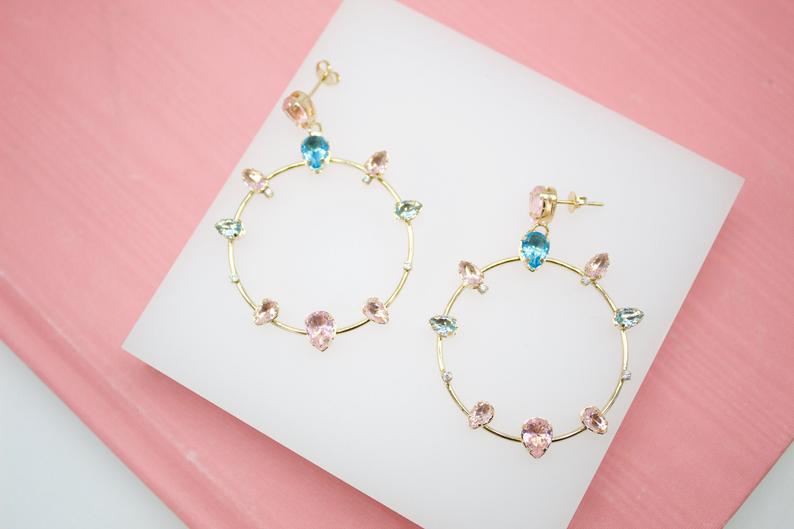 A pair of gold hoop earrings embellished with colorful pink and blue teardrop gemstones rests on a white display against a pink background.