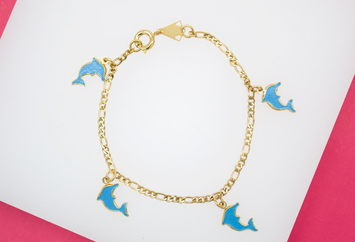 A delicate gold charm bracelet displays four vibrant blue enameled dolphin charms against a white and pink background.
