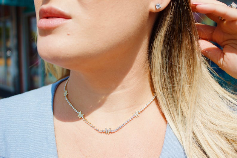 A blonde woman wears a sparkling diamond tennis necklace adorned with gold butterfly charms.