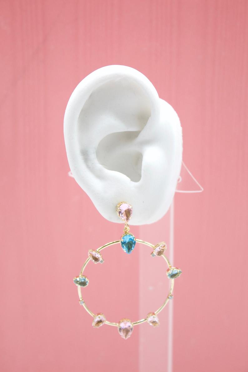 A white mannequin ear displays a gold hoop earring adorned with colorful teardrop crystals against a solid pink background.