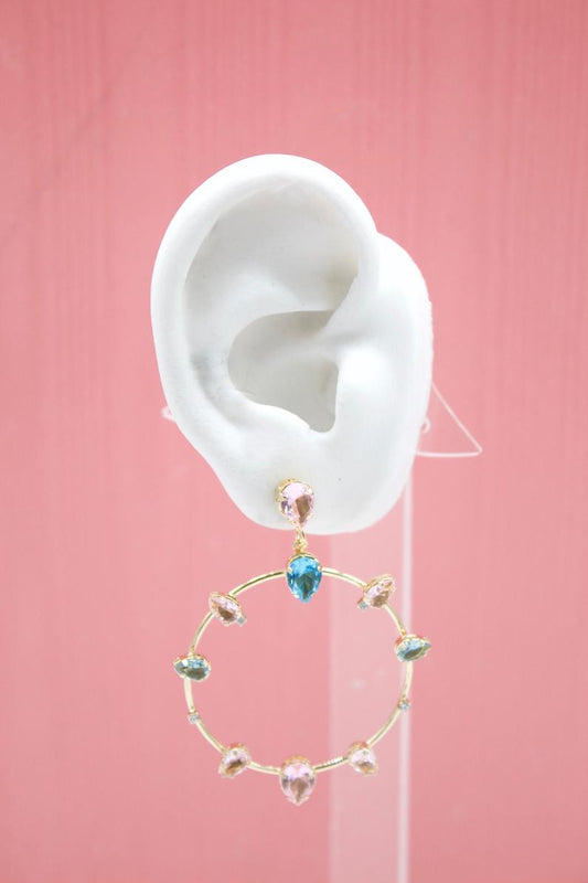 A white mannequin ear displays a gold hoop earring adorned with colorful teardrop crystals against a solid pink background.