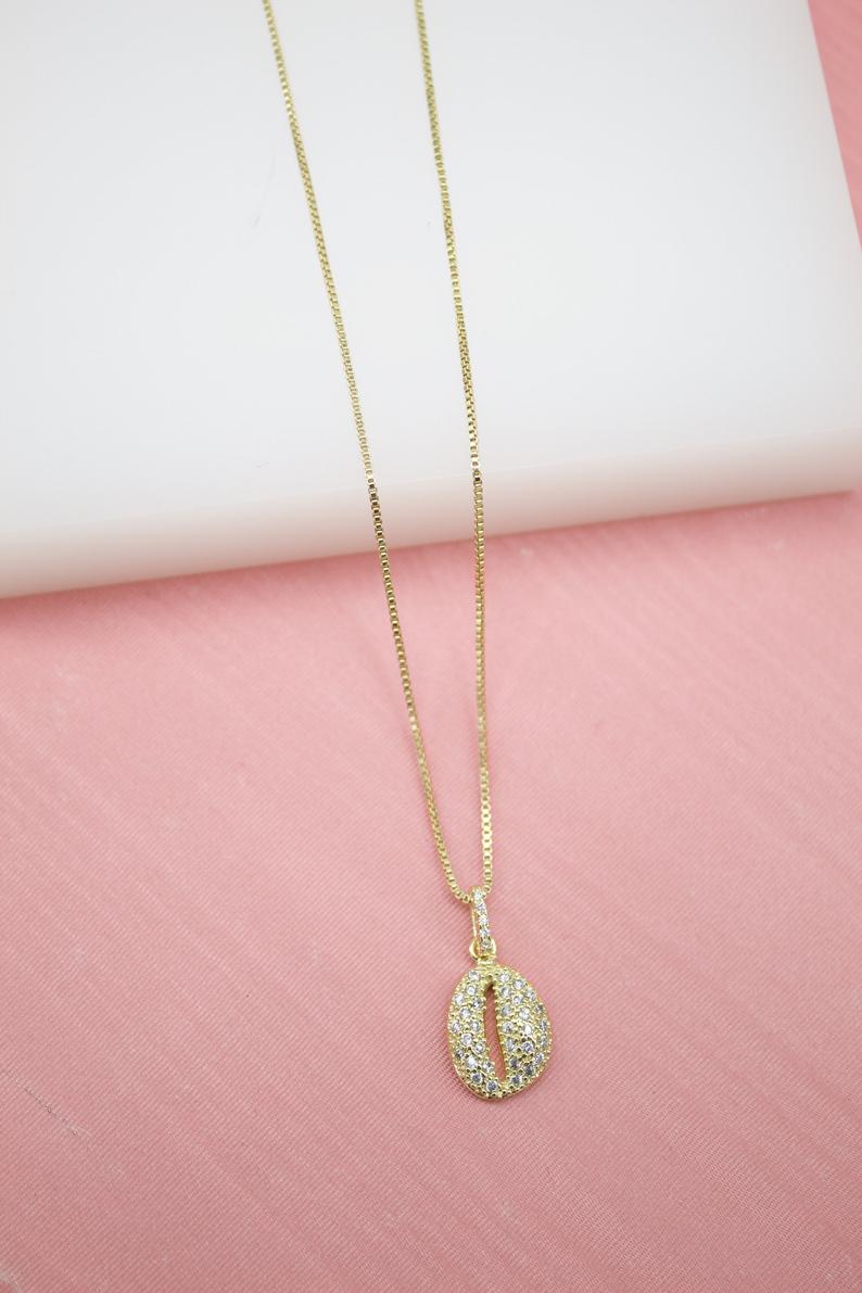 A delicate gold chain holds a sparkling pave cowrie shell pendant against a soft pink and white backdrop.