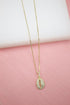 A delicate gold chain holds a sparkling pave cowrie shell pendant against a soft pink and white backdrop.