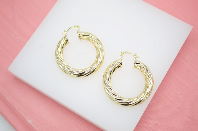 A pair of glossy gold twisted hoop earrings rests on a white display surface against a pink background.
