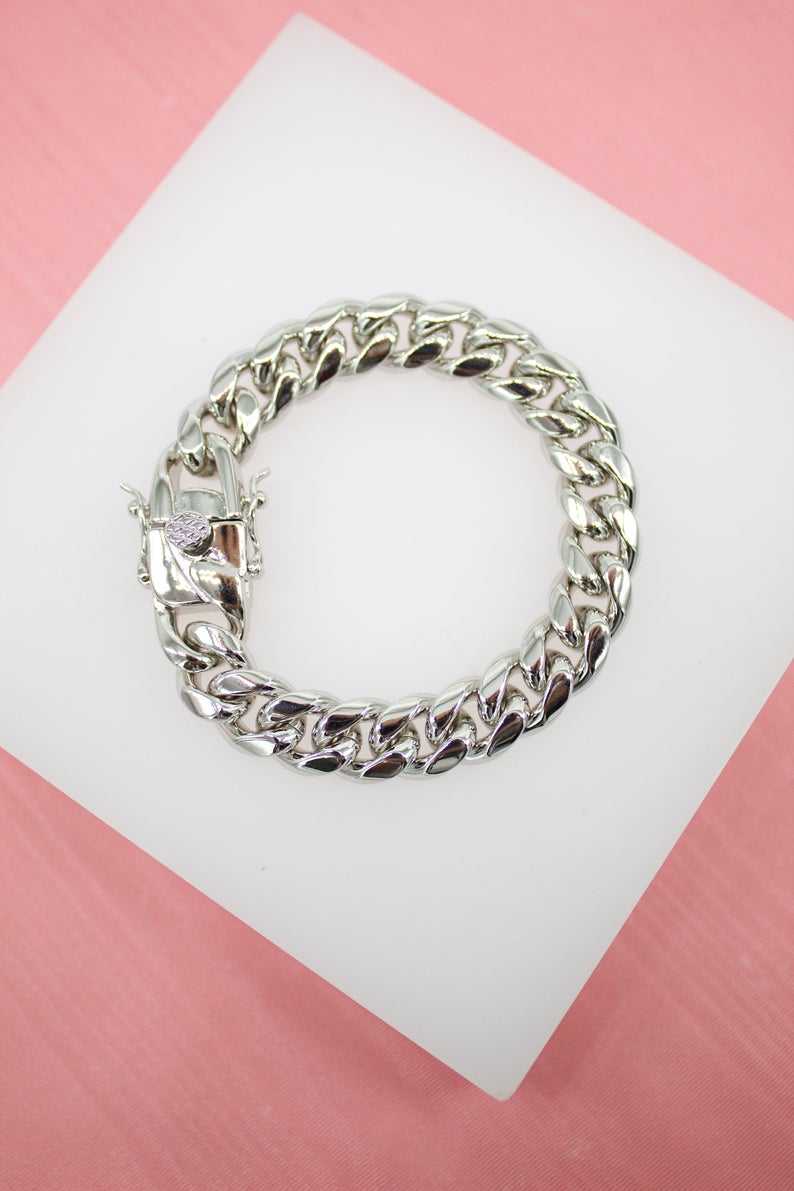 A polished silver cuban link bracelet rests on a white display block against a soft pink background.