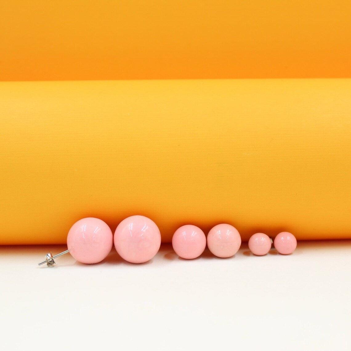 A row of six graduated pink ball stud earrings rests on a white surface against an orange backdrop.