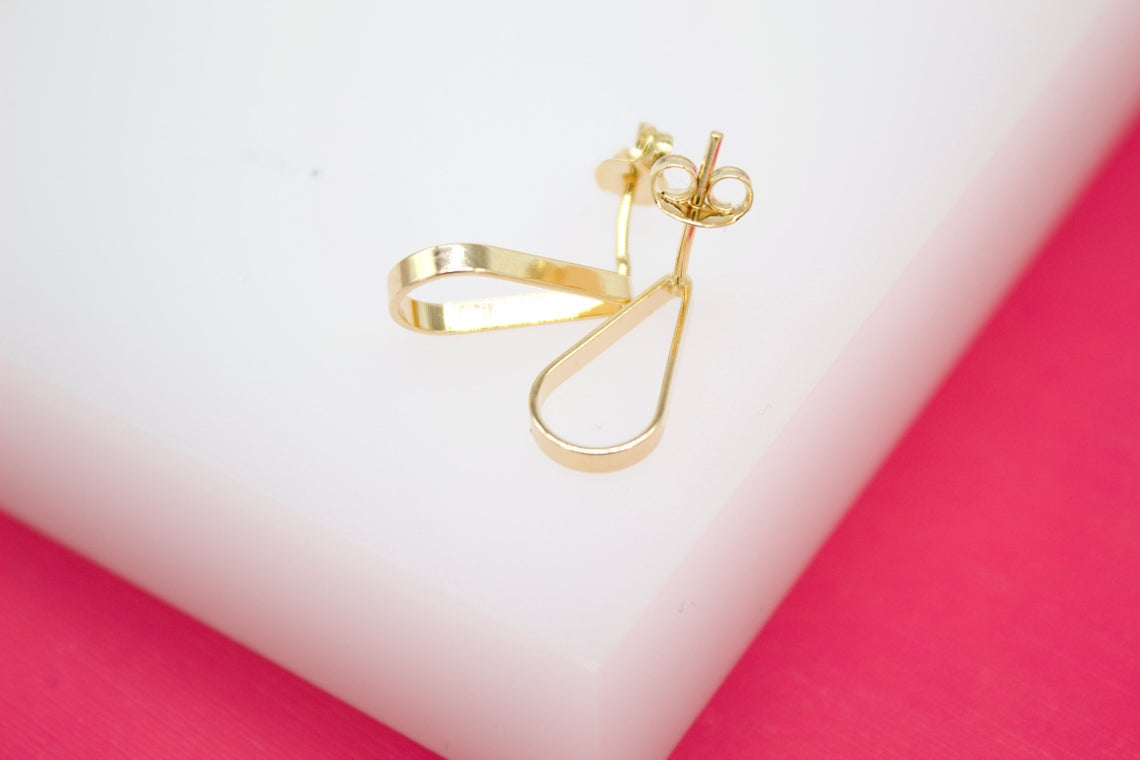 A pair of minimalist gold teardrop earrings rests on a white display stand against a vibrant pink background.