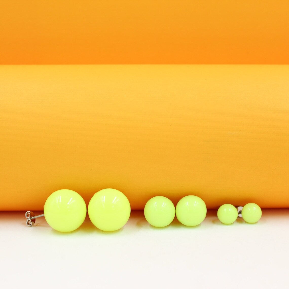 Three pairs of vibrant neon yellow ball stud earrings in graduating sizes sit neatly lined up against a bright orange backdrop.