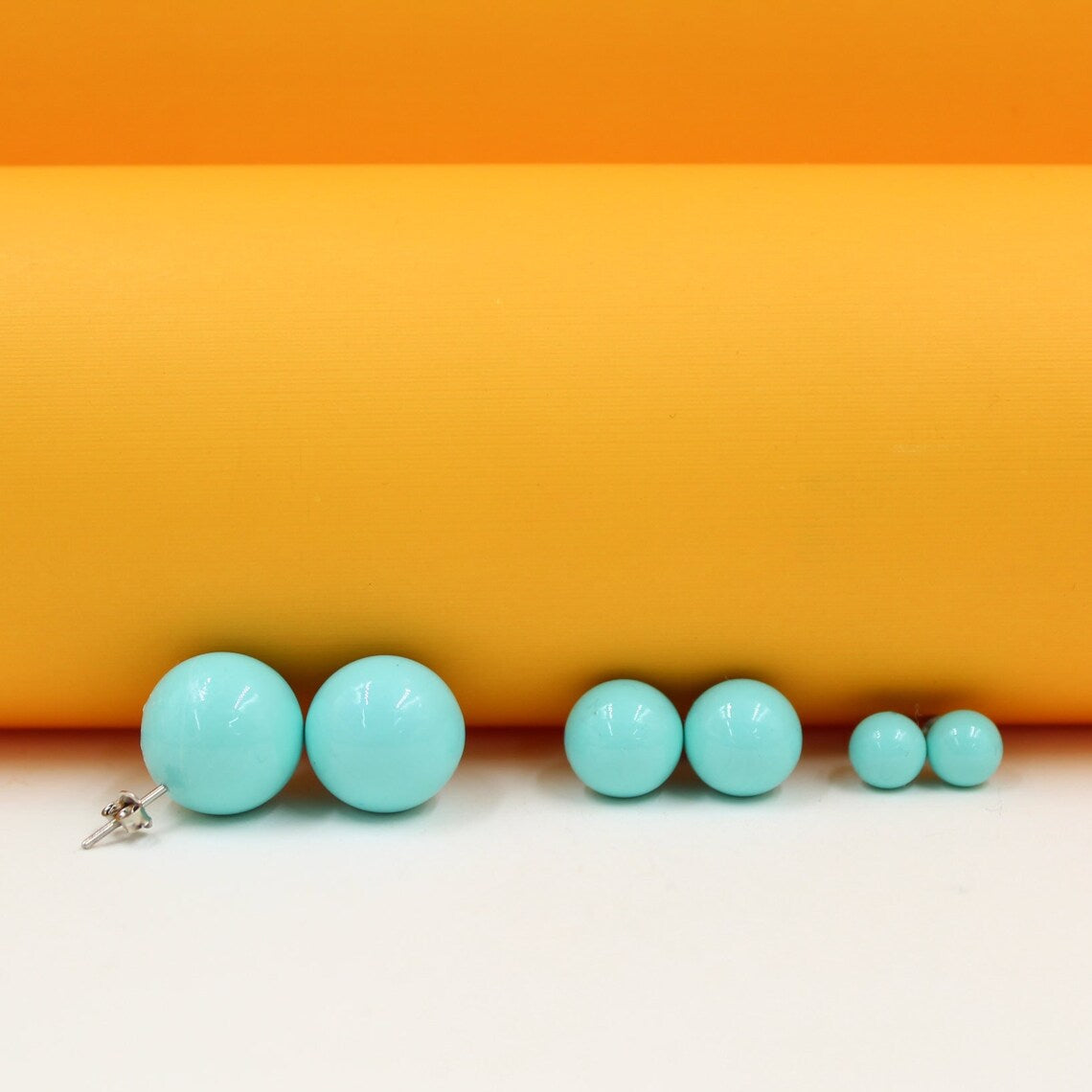 Three pairs of turquoise ball stud earrings in graduating sizes rest against an orange and white backdrop.