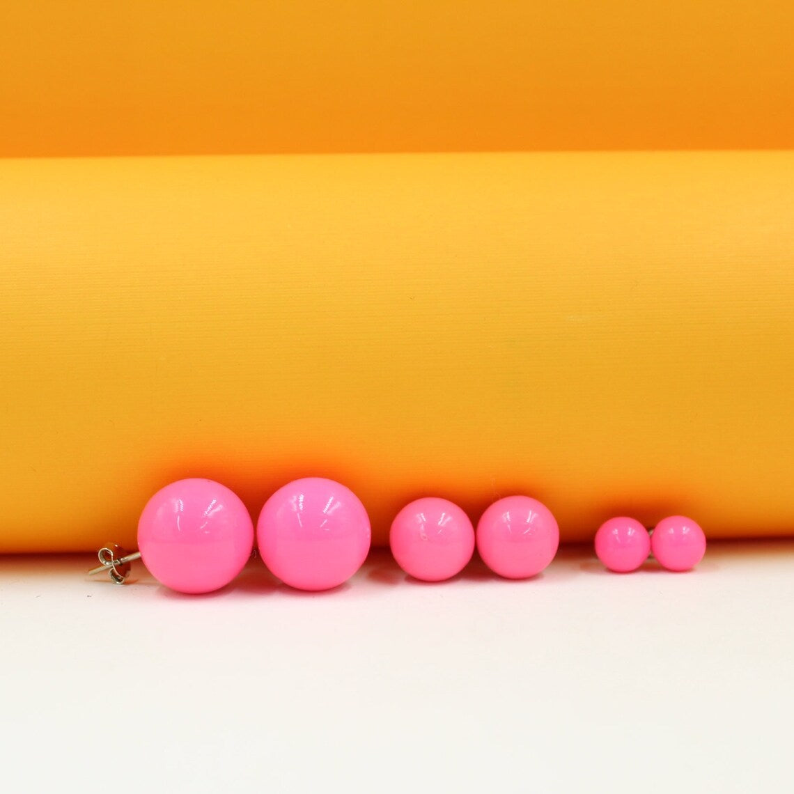 A colorful display of vibrant pink ball stud earrings in graduating sizes rests neatly against a bright orange and white background.