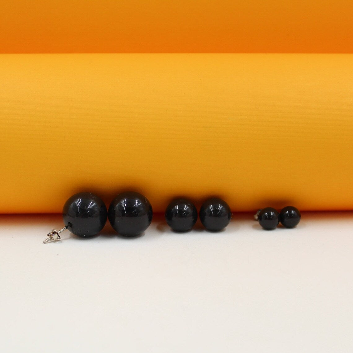 Three pairs of glossy black sphere ball stud earrings sit lined up by size against an orange and white background.