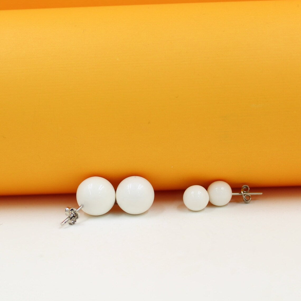 Two pairs of minimalist white ball stud earrings rest on a white surface against a vibrant yellow background.