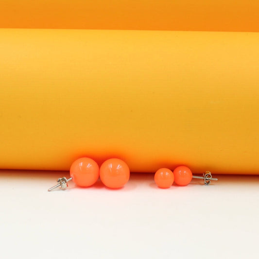 Bright orange ball stud earrings rest on a white surface against a vibrant yellow background.
