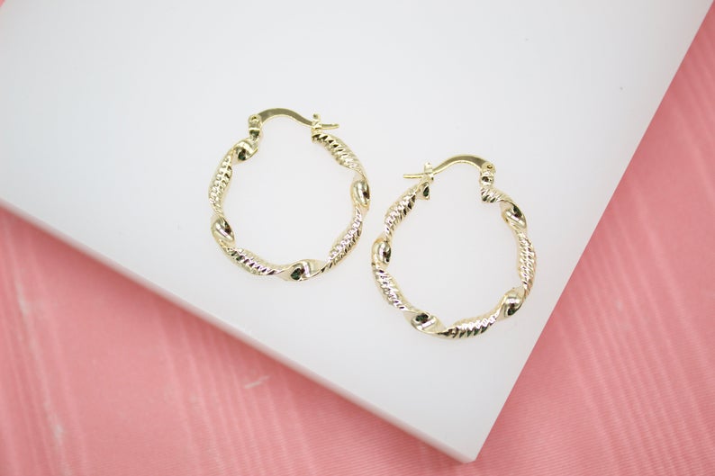 A pair of twisted gold hoop earrings rests on a white display surface against a pink background.