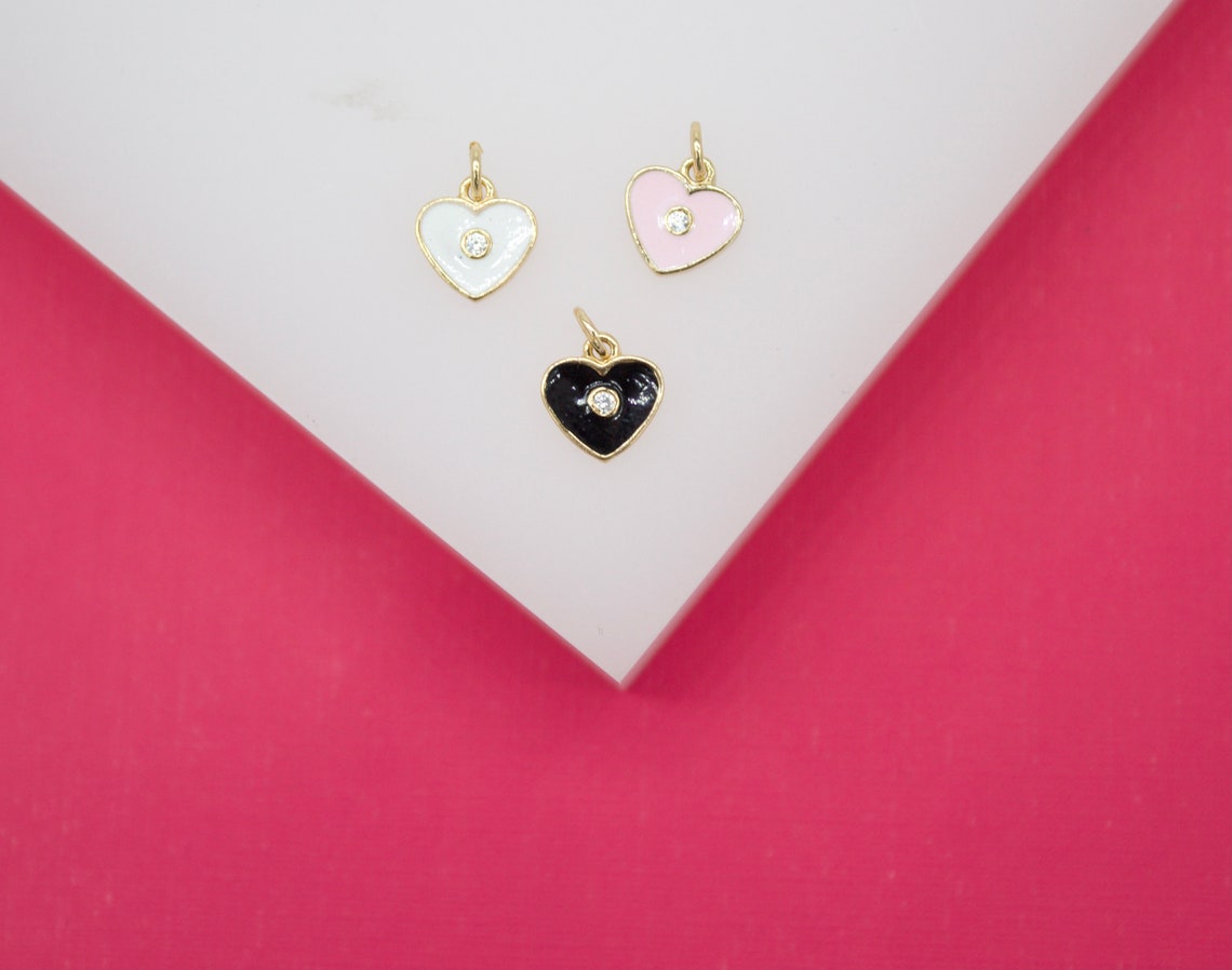 Three gold heart charms featuring white, pink, and black enamel accents rest on a white display surface against a vibrant pink background.