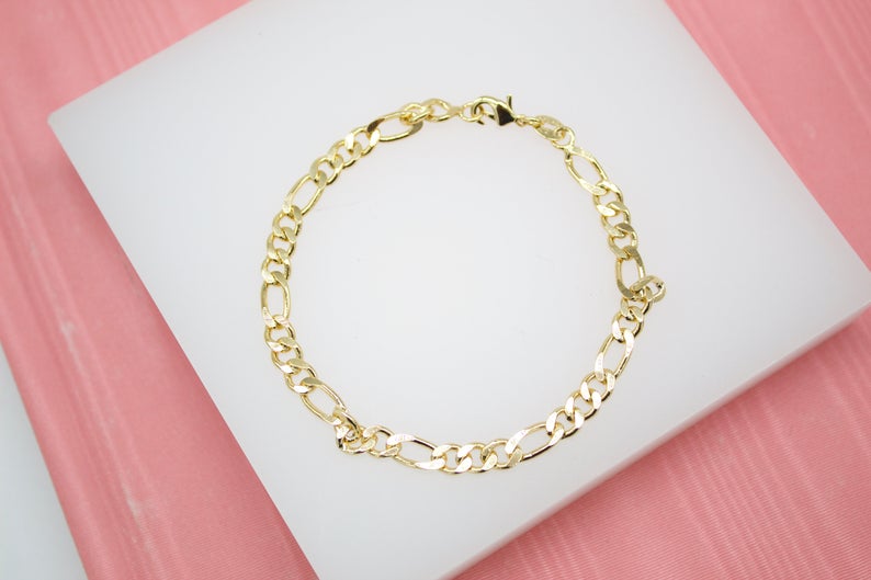 A polished gold Figaro chain bracelet rests neatly on a white display block against a soft pink background.