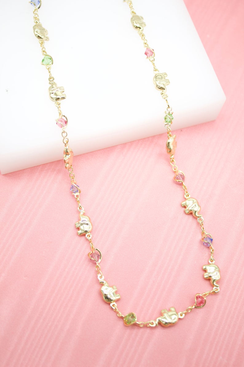 A delicate gold chain necklace features alternating gold elephant charms and colorful crystal beads against a soft pink and white background.