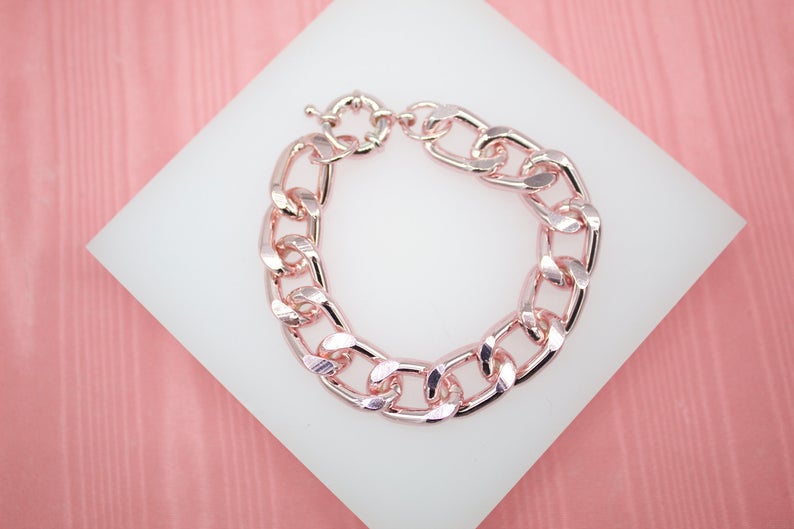 A thick rose gold chain link bracelet rests on a white display pad against a pink background.