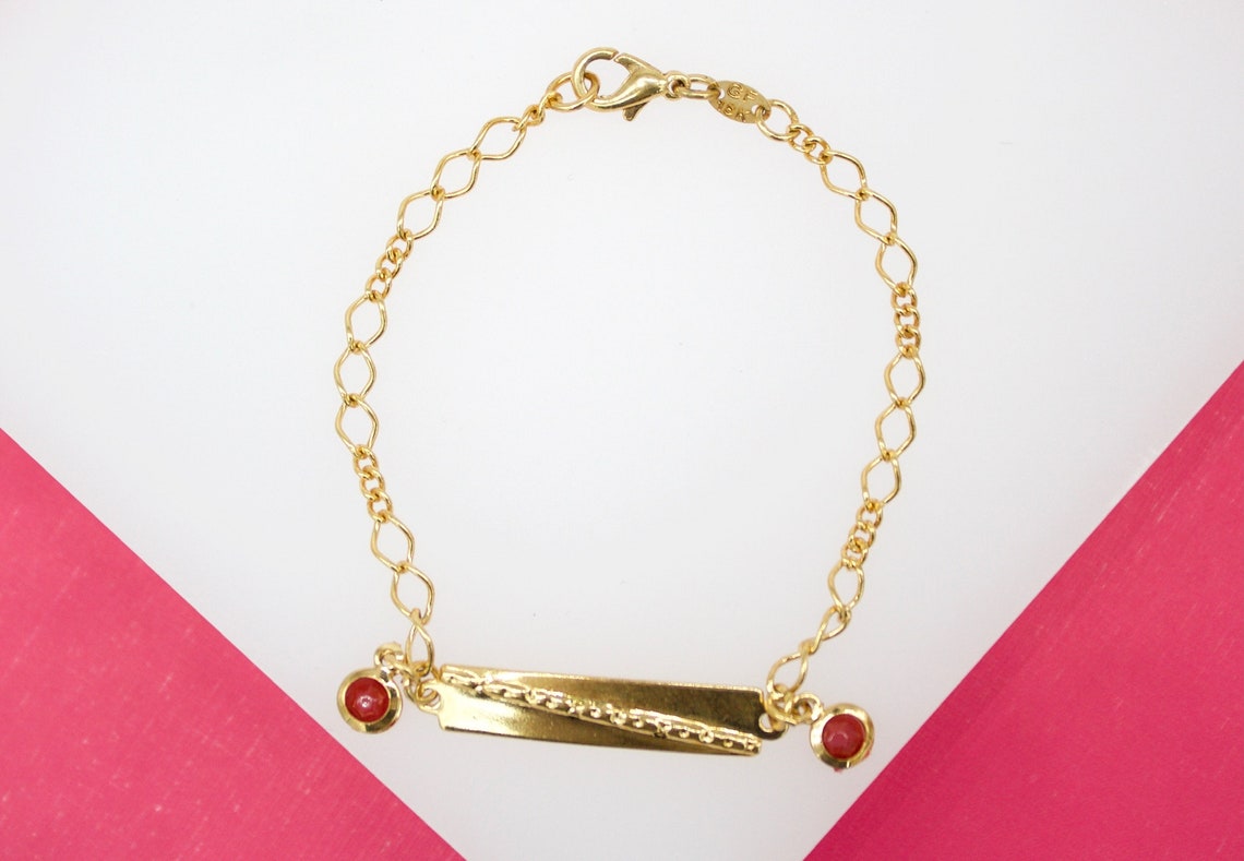 A delicate gold ID bracelet rests on a white and pink background, featuring two small red stone charms flanking a rectangular nameplate.