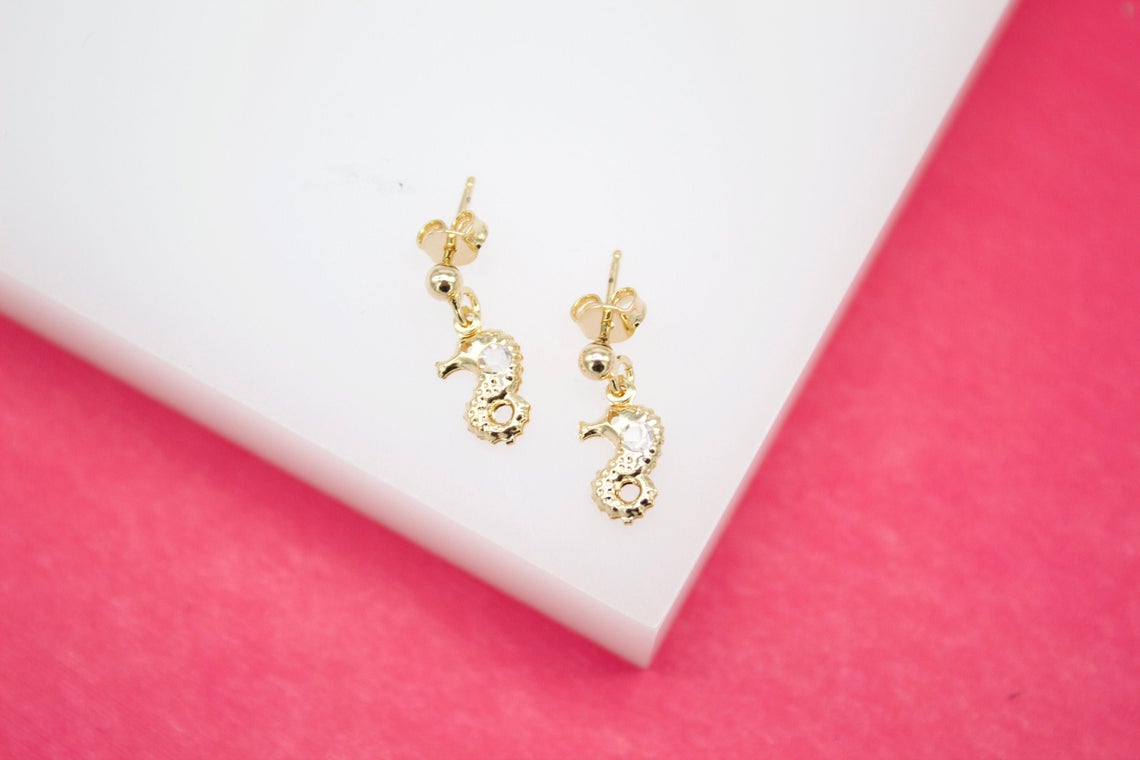 A pair of delicate gold seahorse earrings rest on a white display stand against a vibrant pink background.