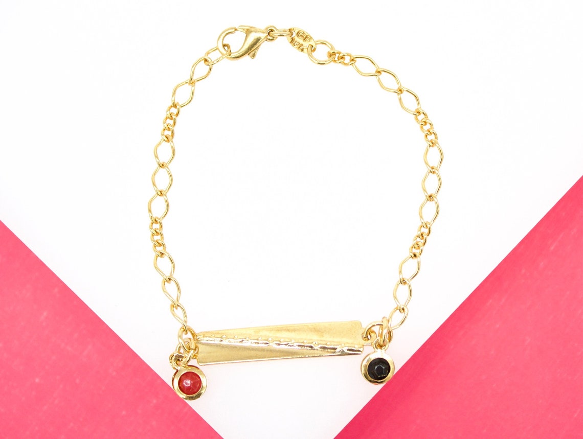 A delicate gold chain bracelet features an engraved bar charm with dangling red and black gemstone pendants against a pink and white background.