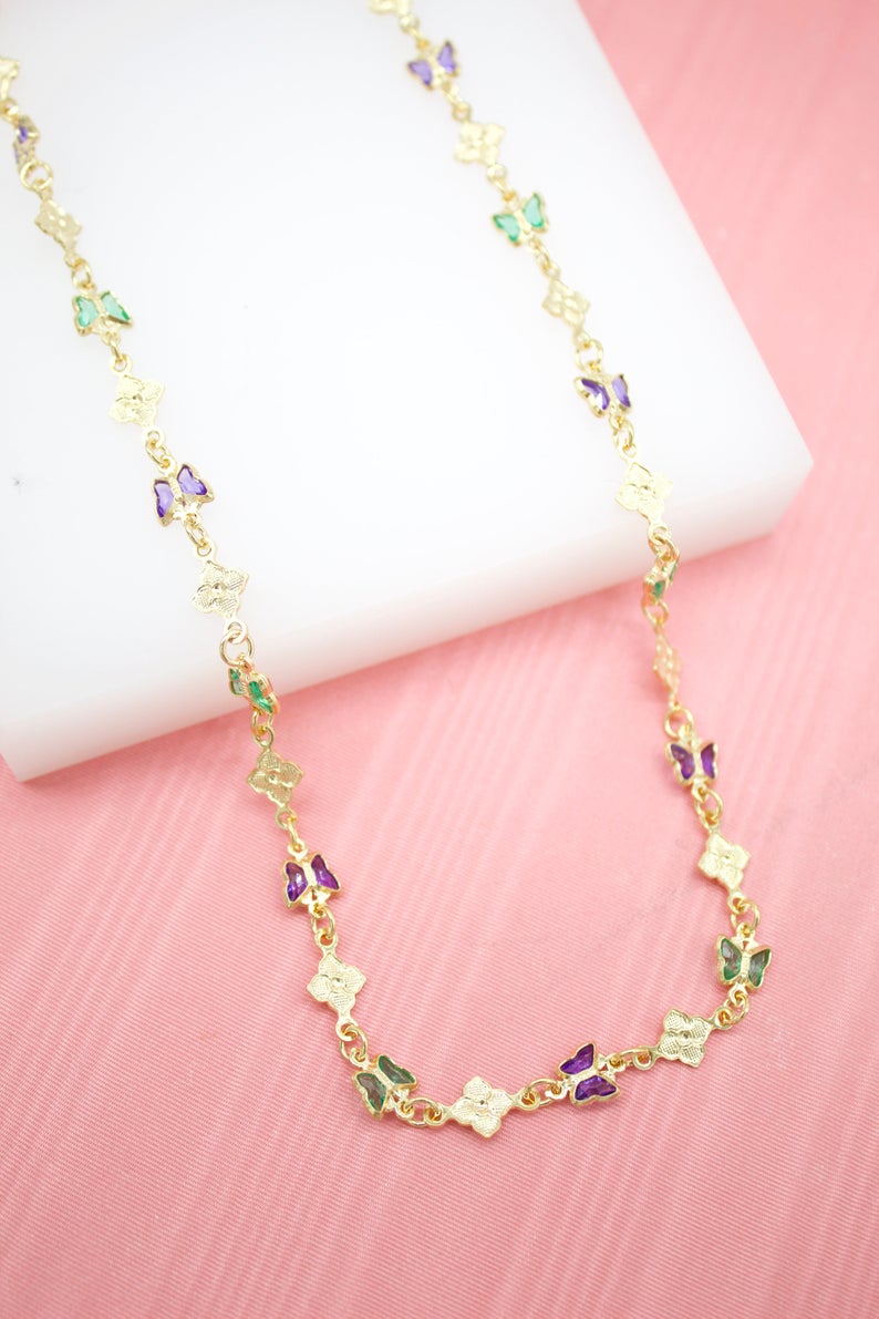 A gold butterfly necklace features alternating purple and green crystal accents along a delicate chain resting on a white display against a pink background.