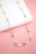 A gold butterfly necklace features alternating purple and green crystal accents along a delicate chain resting on a white display against a pink background.