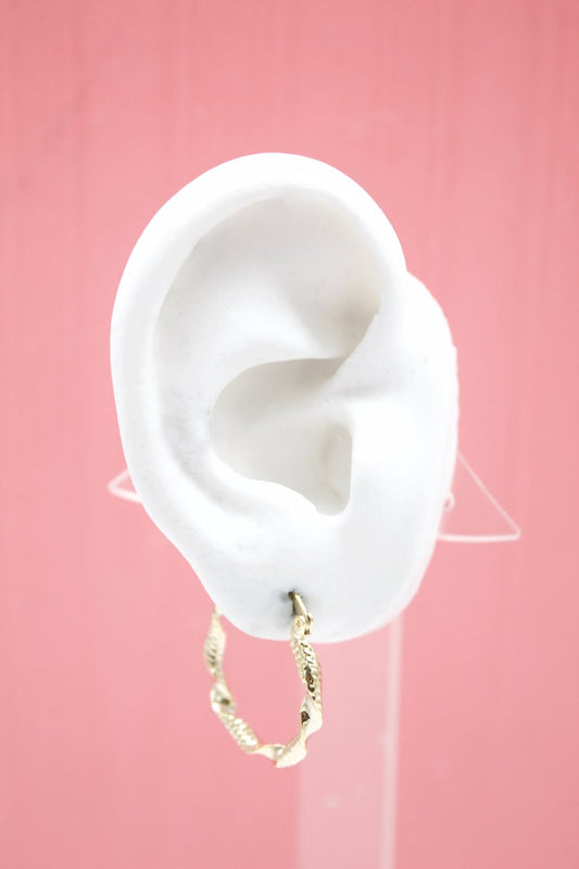 A white mannequin ear displays a twisted gold hoop earring against a solid pink background.
