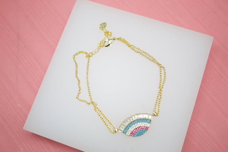 A delicate gold double-strand bracelet featuring a colorful, crystal-embellished evil eye charm rests on a white display surface against a pink background.