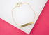A delicate gold ID bracelet rests on a white and pink background, showcasing its shiny link chain and blank metal plaque.