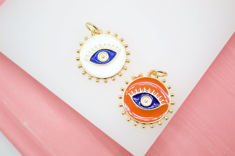Two round gold-plated enamel evil eye charms in white and orange rest on a white display surface against a pink background.
