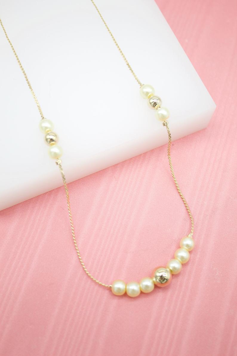 A delicate gold chain necklace features alternating pearls and gold beads resting against a soft pink backdrop.