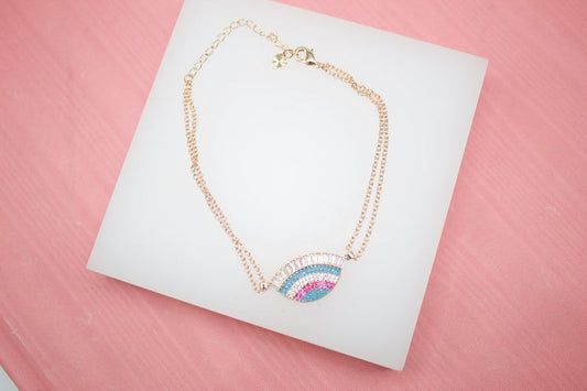 A delicate rose gold double-chain anklet rests on a white display block, featuring an eye-shaped charm embellished with colorful cubic zirconia stones.