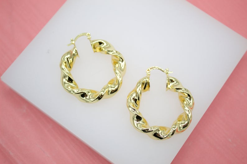 A pair of polished gold twist hoop earrings rests on a white display surface against a pink background.