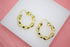 A pair of polished gold twist hoop earrings rests on a white display surface against a pink background.