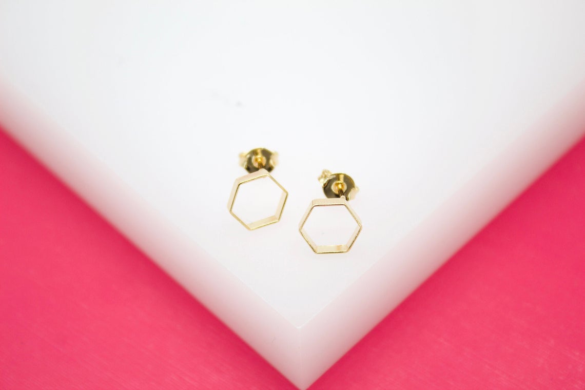 Two gold geometric hexagon stud earrings rest on a white display surface against a vibrant pink background.