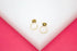 Two gold geometric hexagon stud earrings rest on a white display surface against a vibrant pink background.