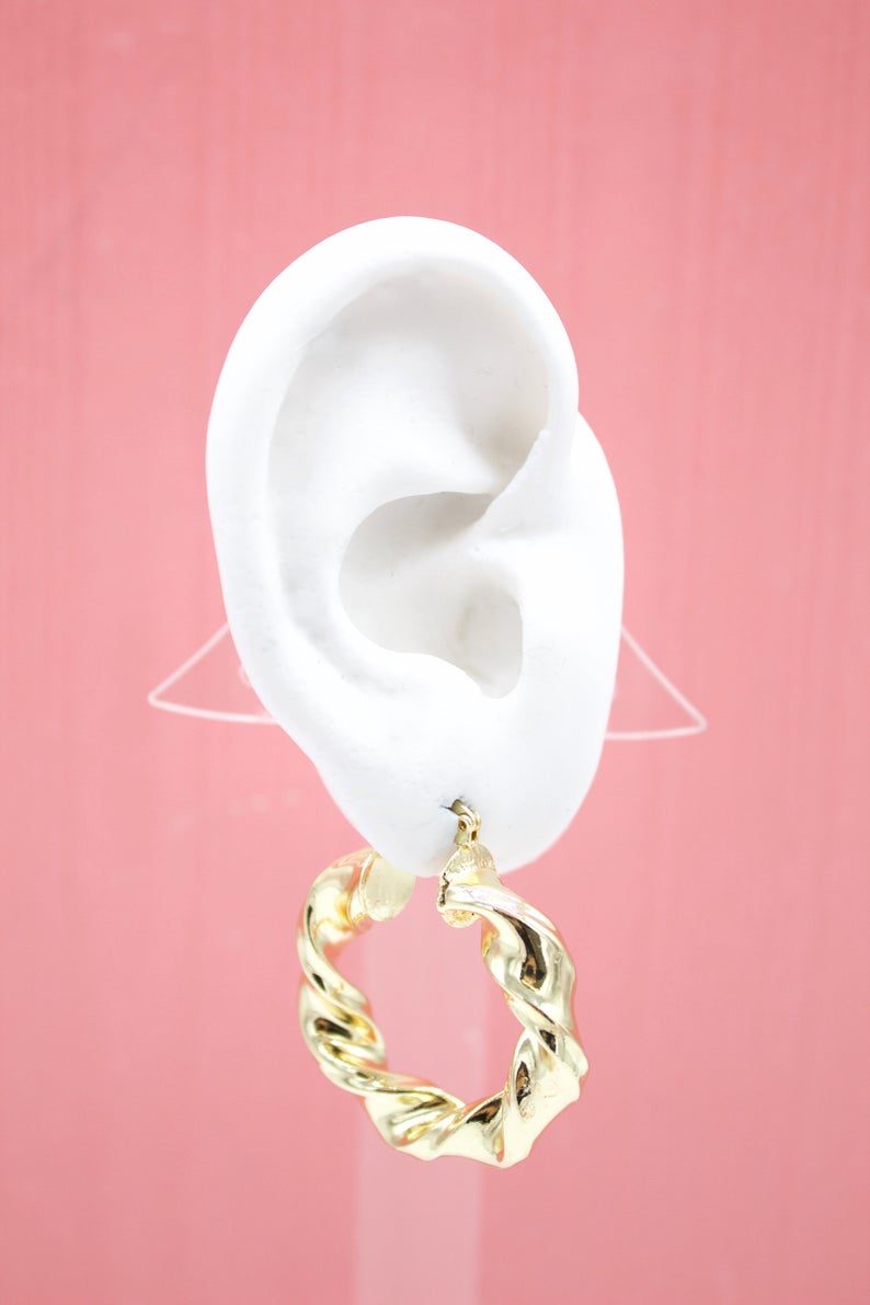 A white mannequin ear displays a shiny gold twisted hoop earring against a solid pink backdrop.