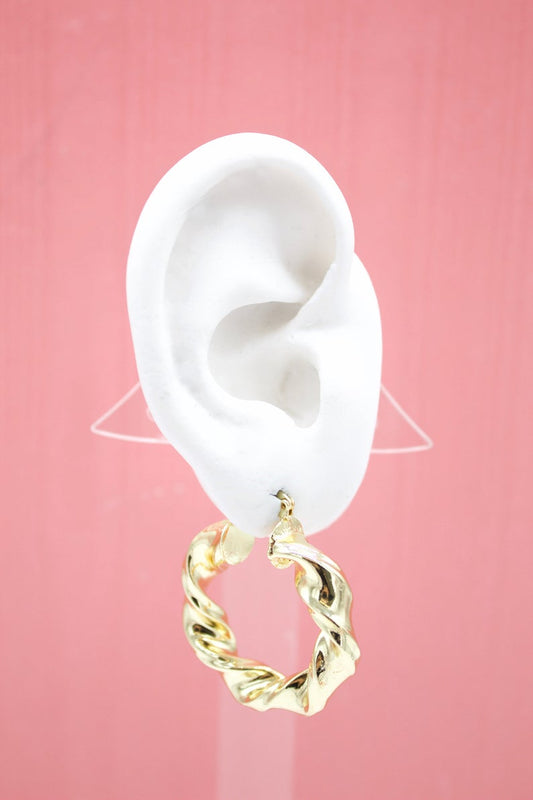 A white mannequin ear displays a shiny gold twisted hoop earring against a solid pink backdrop.