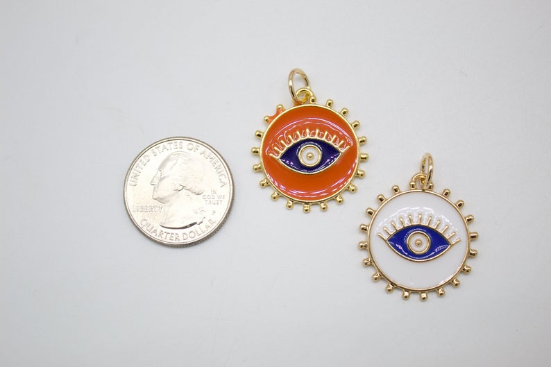 An orange and a white enamel evil eye charm sit next to a US quarter coin for scale.