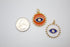 An orange and a white enamel evil eye charm sit next to a US quarter coin for scale.