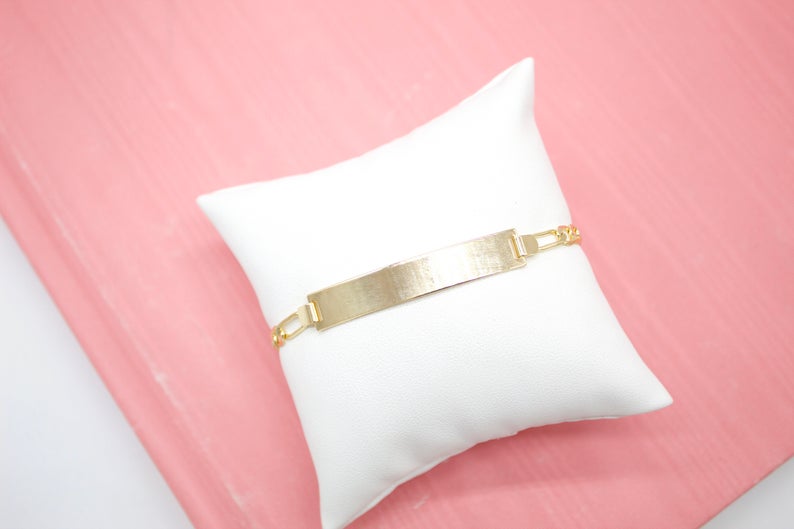 A gold identification bracelet rests on a small white display pillow against a pink background.