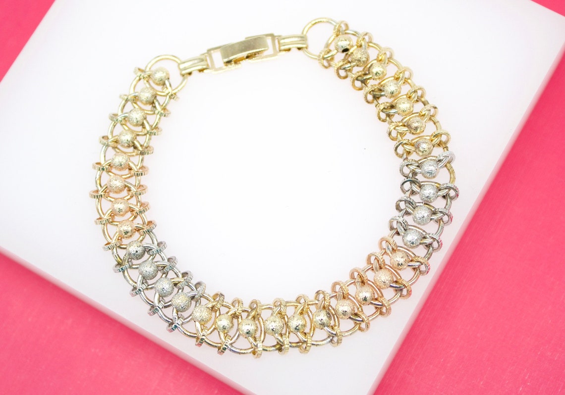 A tri-color gold multi-strand link bracelet featuring textured beaded details rests on a white display against a pink background.
