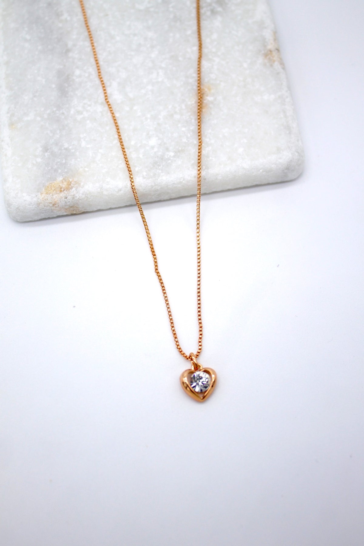A delicate gold heart pendant necklace featuring a sparkling diamond accent rests elegantly against a white marble display.