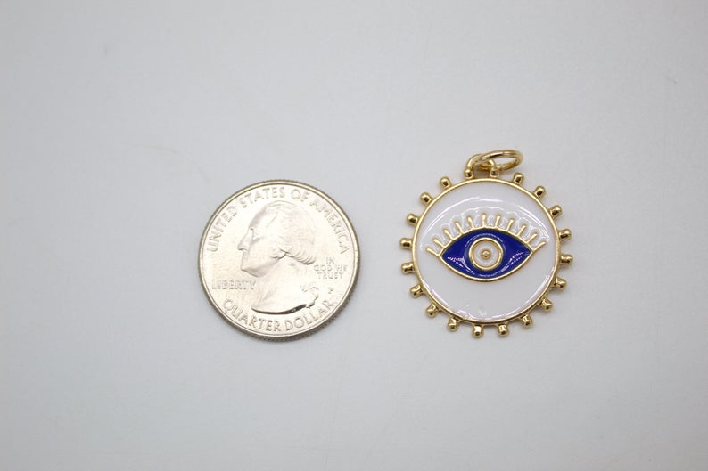 A round, gold-rimmed evil eye pendant with blue and white enamel rests beside a US quarter for scale.