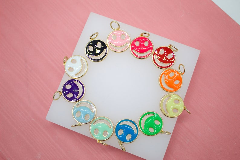 A colorful assortment of enamel smiley face charms with gold trim rests on a white display against a pink background.
