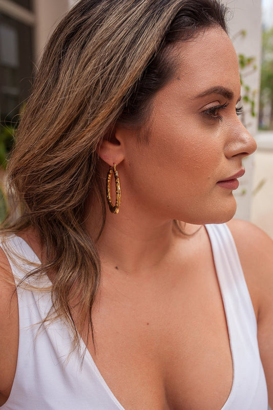 A young woman wearing stylish gold bamboo hoop earrings looks away while posing outdoors in a white top.